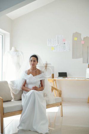 Foto de Novia pensativa en hermoso vestido de novia y velo sentado en el taller de moda - Imagen libre de derechos