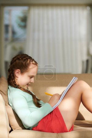 Téléchargez les photos : Souriant preteen fille assis sur canapé et faire des devoirs - en image libre de droit