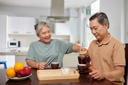 Téléchargez les photos : Femme âgée versant le thé dans la tasse de son mari au petit déjeuner - en image libre de droit