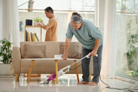 Foto de Mujer mayor piso de limpieza de vacío cuando el marido limpiar el polvo - Imagen libre de derechos