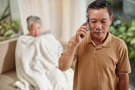 Photo for Serious senior man talking on phone, describing symptoms to doctor, his sick wife is sitting in background - Royalty Free Image