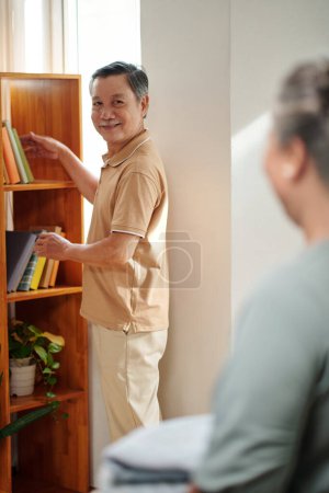 Foto de Sonriente hombre mayor poniendo libros en el estante y mirando a la esposa - Imagen libre de derechos