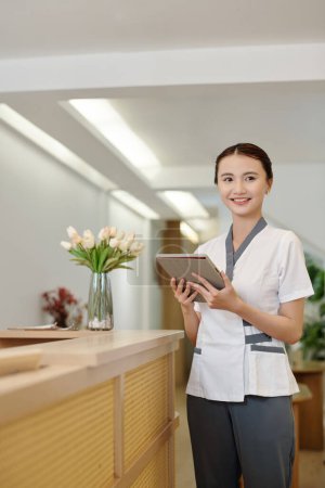 Téléchargez les photos : Joyeuse réceptionniste de salon de spa vérifiant l'horaire sur tablette avant l'ouverture - en image libre de droit
