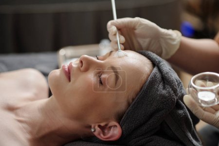 Kosmetikerin trägt Glykolsäure-Peeling auf Gesicht junger Frau auf