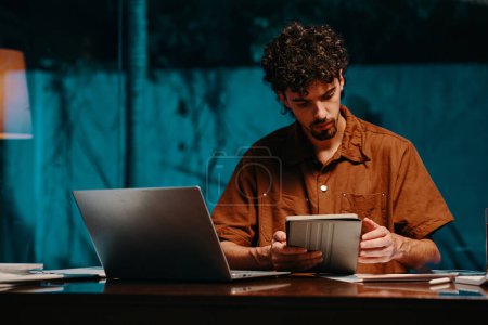 Téléchargez les photos : Homme d'affaires sérieux assis au bureau et lisant le rapport sur tablette tard dans la nuit - en image libre de droit
