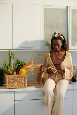 Foto de Mujer joven negra sentada en el mostrador de kichen y pelando fruta de la pasión - Imagen libre de derechos