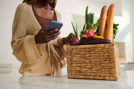 Photo for Woman checking basket of fresh fruits and vegetables she ordered online via mobile app - Royalty Free Image