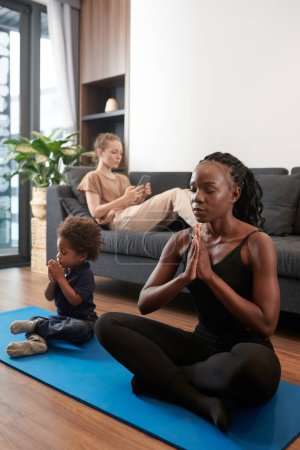 Foto de Mujer joven y su hijo pequeño meditando sentados en posición de loto en la esterilla de yoga en casa - Imagen libre de derechos