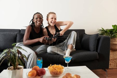 Foto de Emocionada joven pareja lesbiana diversa comiendo bocadillos, bebiendo cócteles y jugando videojuegos en casa - Imagen libre de derechos