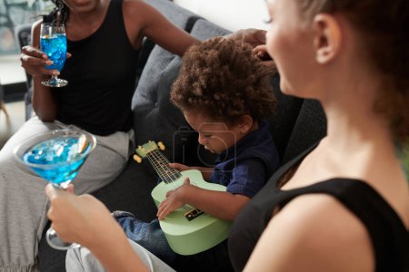 Foto de Niño jugando ukelele cuando sus madres hablando y bebiendo cócteles en casa - Imagen libre de derechos