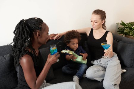 Foto de Diverso joven pareja bebiendo cócteles de frutas cuando pequeño hijo jugando ukelele - Imagen libre de derechos
