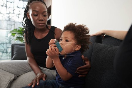 Téléchargez les photos : Mère inquiète regardant son fils utiliser un inhalateur lors d'une crise d'asthme à la maison - en image libre de droit