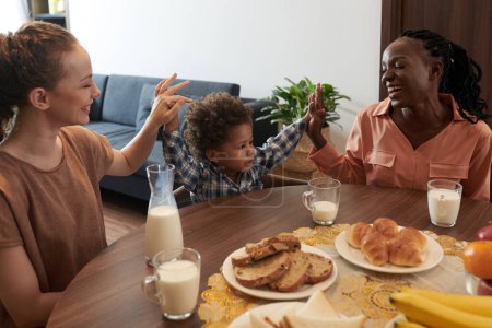 Foto de Feliz diversas madres dando alta cinco a su pequeño hijo cuando se sienta en la mesa de la cocina con taza de leche - Imagen libre de derechos