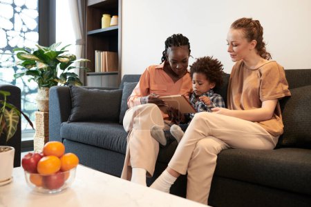 Téléchargez les photos : Mères et fils assis sur le canapé à la maison et jouer au jeu sur tablette - en image libre de droit