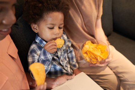 Téléchargez les photos : Tout-petit garçon manger des chips de légumes en regardant dessin animé sur ordinateur portable avec sa famille - en image libre de droit
