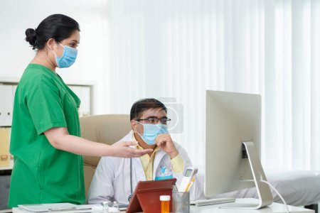 Photo for Surgeon and physician in protective masks discussing medical history of patient on computer screen - Royalty Free Image
