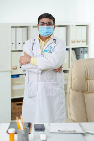 Photo for Portrait of serious Indian general practitioner in medical mask crossing arms when standing at desk in his office - Royalty Free Image