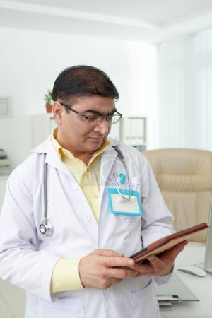 Téléchargez les photos : Médecin généraliste fronçant les sourcils lisant les antécédents médicaux du patient sur tablette - en image libre de droit