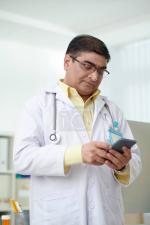 Téléchargez les photos : Médecin généraliste indien mature sérieux lisant des messages texte sur smartph - en image libre de droit