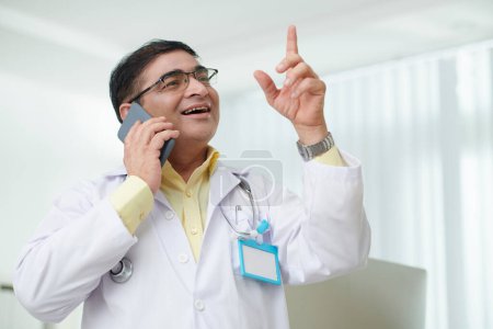 Foto de Retrato de médico general maduro excitado hablando en ph con paciente o compañero de trabajo de otro hospital - Imagen libre de derechos