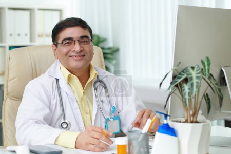 Photo for Portrait of smiling mature general practitioner in glasses sitting at desk in his office - Royalty Free Image
