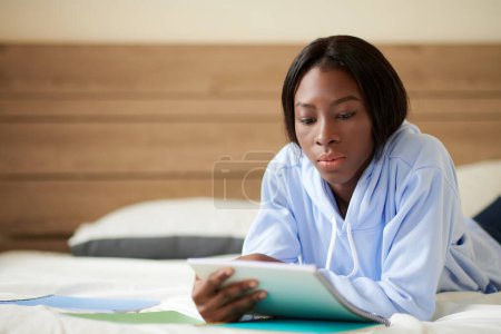 Téléchargez les photos : Pensif jeune femme noire couchée sur le lit et la lecture de notes dans le carnet lors de la préparation à l'examen - en image libre de droit
