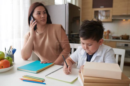 Téléchargez les photos : Écolier faire des devoirs sous le contrôle de sa mère parler au téléphone avec enseignant - en image libre de droit