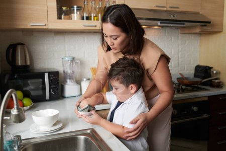 Photo for Young woman explaining son how to wipe dishes after cleaning - Royalty Free Image