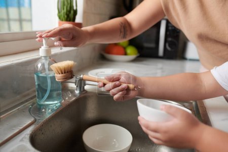 Foto de Las manos de la madre ayudando a su hijo a agregar detergente lavavajillas en el cepillo cuando está limpiando cuencos después del desayuno - Imagen libre de derechos
