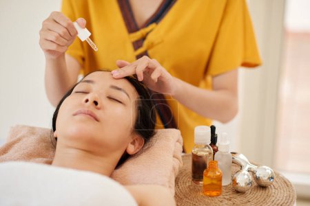 Foto de Beauticial aplicando suero rejuvenecedor en la piel de cliente femenina relajante antes de darle su masaje facial - Imagen libre de derechos