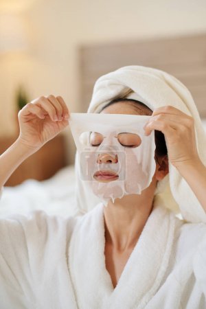 Téléchargez les photos : Calme jeune femme appliquant masque feuille de refroidissement sur le visage le matin - en image libre de droit