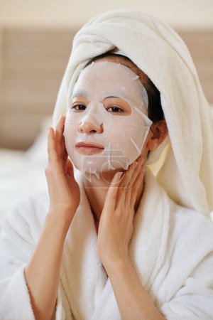 Photo for Portrait of positve young woman applying soothing and hydrating sheet mask on face after morning shower - Royalty Free Image