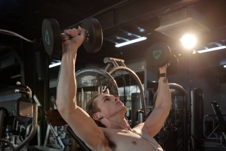 Photo for Fit young man listening to music in earbuds when doing dumbbell press on bench - Royalty Free Image
