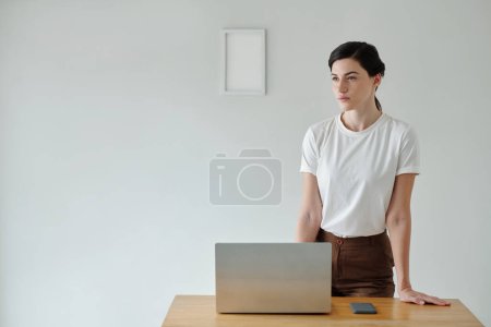 Téléchargez les photos : Pensive jeune femme debout à la table avec ordinateur portable ouvert - en image libre de droit