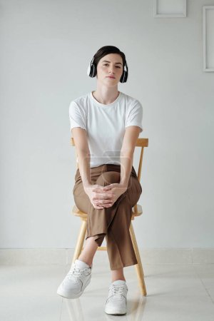 Foto de Mujer joven sentada en silla y escuchando música en auriculares - Imagen libre de derechos