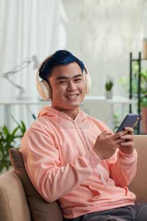 Photo for Joyful young man sitting on couch and listening to music from playlist on his smartphone - Royalty Free Image