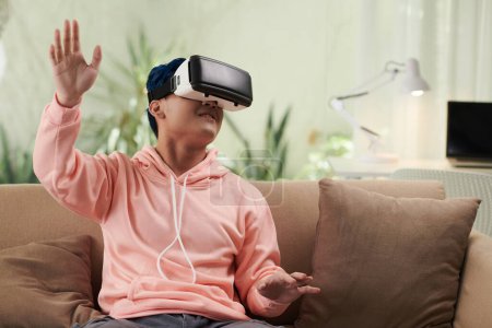 Foto de Joven alegre jugando videojuego en gafas de realidad virtual cuando pasa el fin de semana en casa - Imagen libre de derechos