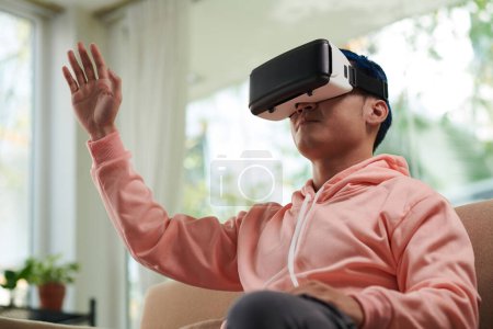Téléchargez les photos : Jeune homme explorant métaverse assis sur le canapé à la maison dans un casque VR - en image libre de droit