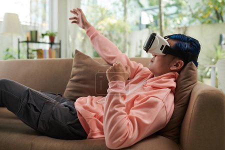 Photo for Zoomer lying on couch at home and playing videogame in vr glasses - Royalty Free Image
