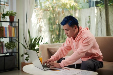 Téléchargez les photos : Sérieux jeune designer UI avec les cheveux bleus travaillant sur ordinateur portable à la maison - en image libre de droit