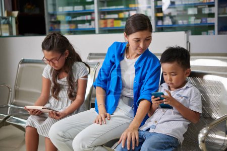 Téléchargez les photos : Mère et deux enfants attendent dans le hall de l'hôpital, regardant des vidéos sur smartphone et tablette - en image libre de droit