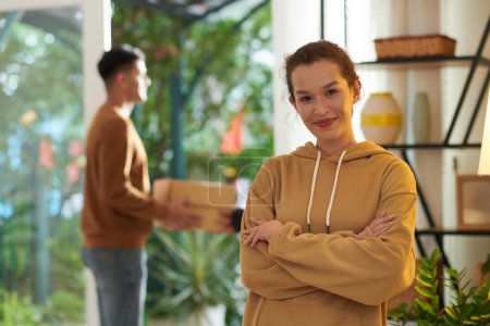 Photo for Portrait of happy confident young woman standing with arms crossed, her boyfriend receiving parcel in background - Royalty Free Image