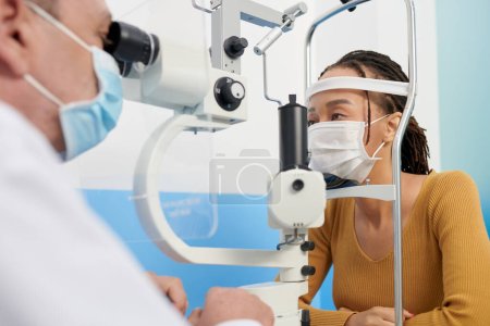 Foto de Mujer negra en máscara médica obtener ojos examinados con autorefractómetro para obtener la prescripción de lentes - Imagen libre de derechos
