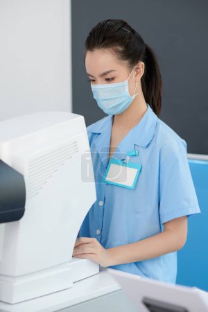 Photo for Portrait of young ophthalmic nurse checking information on auto refractometer when getting objective measurement of patients refractive error - Royalty Free Image