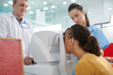 Photo for Female patient putting head in autorefractor when doctor and medical nurse checking her eyes - Royalty Free Image