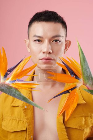 Foto de Retrato de un hombre joven con delineador de ojos y pedrería maquillaje al estar de pie entre las flores strelitzia - Imagen libre de derechos