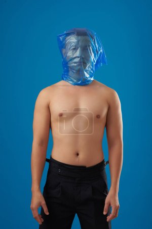 Photo for Portrait of serious shirtless young man standing with blue plastic bag on his head, ecology concept - Royalty Free Image