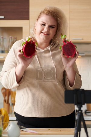Photo for Smiling Plus-size young woman showing cut dragon fruit during live stream with subscribers - Royalty Free Image