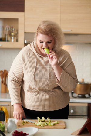 Photo for Curvy young woman tasting green apple she is cutting for fruit salad at kitchen table - Royalty Free Image
