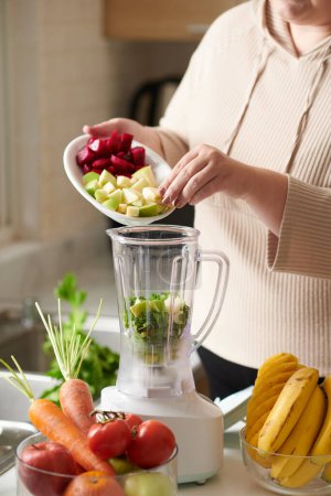 Photo for Plus-size woman putting cut celery, apple and dragon fruit in blender when making morning smoothie - Royalty Free Image